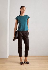 Teal kortermet t-skjorte med blonder, sammen med svarte smale bukser og svarte hælmuler, holdt en svart veske. Bakgrunn med lys tregulv.
