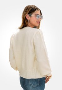 Mujer con un suéter de punto crema y mangas abullonadas, con jeans azules, girada ligeramente hacia un lado y usando unas grandes gafas de sol.