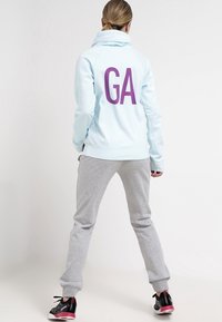 Sudadera azul clara con un estampado morado de "GA" en la parte posterior, combinada con pantalones deportivos grises. La sudadera presenta un cuello alto y puños acanalados.