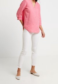 Camicia lunga a maniche con scollo a V in lino rosa, con dettagli di bottoni, abbinata a jeans skinny bianchi e scarpe slip-on bianche con un motivo in rete.