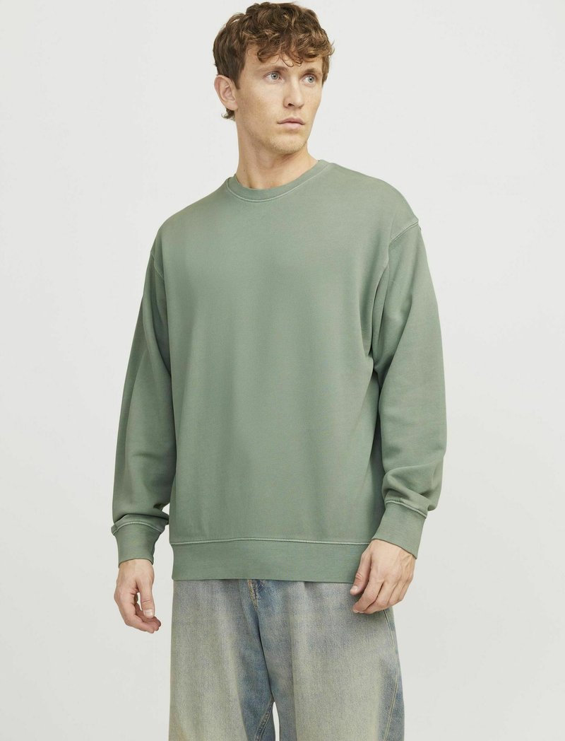 Felpa verde in cotone con collo rotondo e maniche lunghe, caratterizzata da una vestibilità rilassata e polsini a coste, abbinata a jeans azzurri chiari.