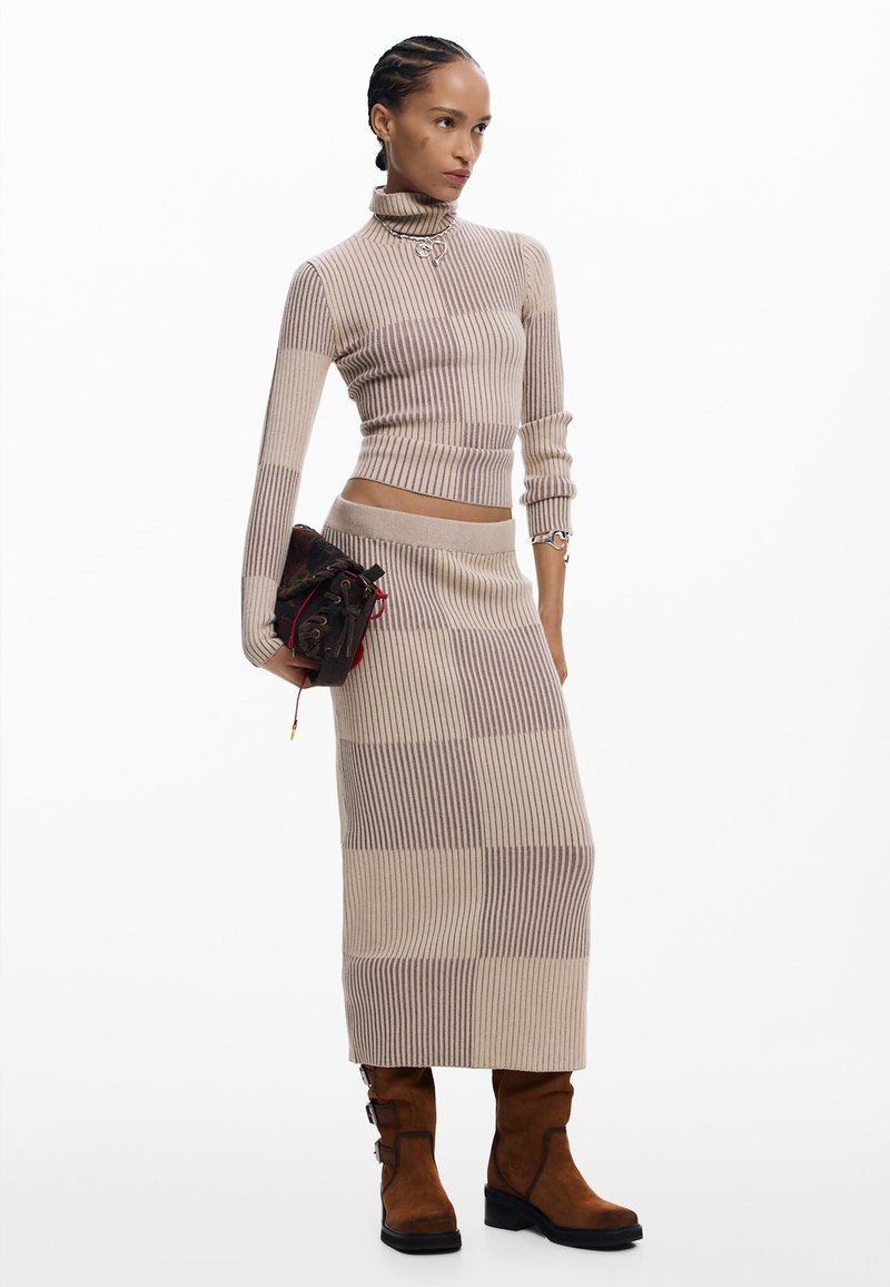 Woman stands wearing beige and taupe ribbed knit turtleneck and matching long skirt, holding dark patterned clutch and brown mid-calf boots.