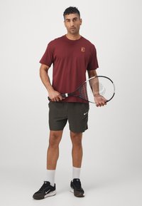 Burgundin lyhythihainen t-paita, jossa on logo, oliivinvihreät shortsit, mustat urheilujalkineet ja valkoiset nilkkasukat. Pitää mustaa tennismailaa.