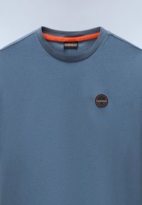 Modrá bavlněná tričko s kulatým výstřihem. Na hrudi má malé kulaté logo v černé a oranžové barvě. Hladká textura, jednoduchý design.