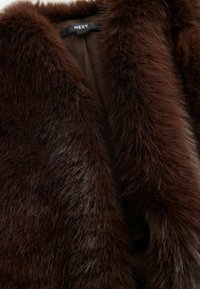 Étole en fausse fourrure marron avec une texture douce et pelucheuse. Le design présente une profonde encolure en V et une doublure intérieure lisse. Etiquette de la marque visible.