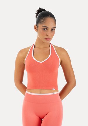 Młoda kobieta z włosami związanymi do tyłu, ubrana w koralowy top na ramiączkach i dopasowane legginsy z wysokim stanem, stojąca z rękami za plecami.