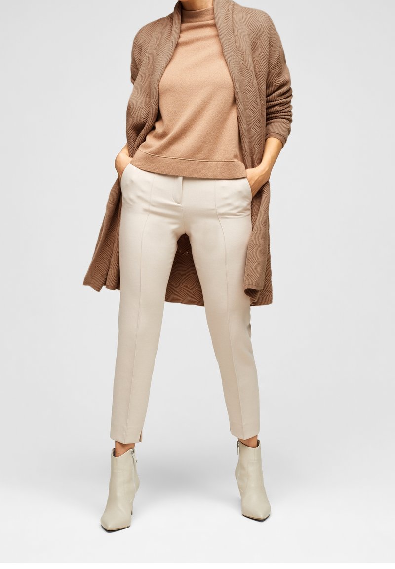 Femme portant un pull à col roulé beige, un long cardigan marron, un pantalon crème ajusté et des bottines crème avec détail fermeture éclair.