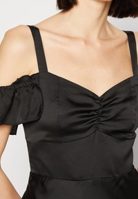 Černé saténové šaty s přiléhavým živůtkem, širokými ramínky a volánkovými detaily odhalených ramen. Nařasený střed a hladká textura.