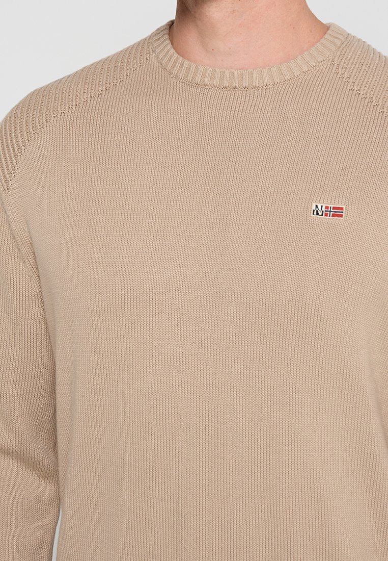 Mężczyzna w beżowym swetrze dzianinowym z okrągłym dekoltem i małym logo norweskiej flagi na klatce piersiowej.