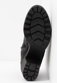 Semelle d'une botte noire avec des crampons profonds appuyée verticalement contre une surface blanche, indiquant la pointure 37 au talon.