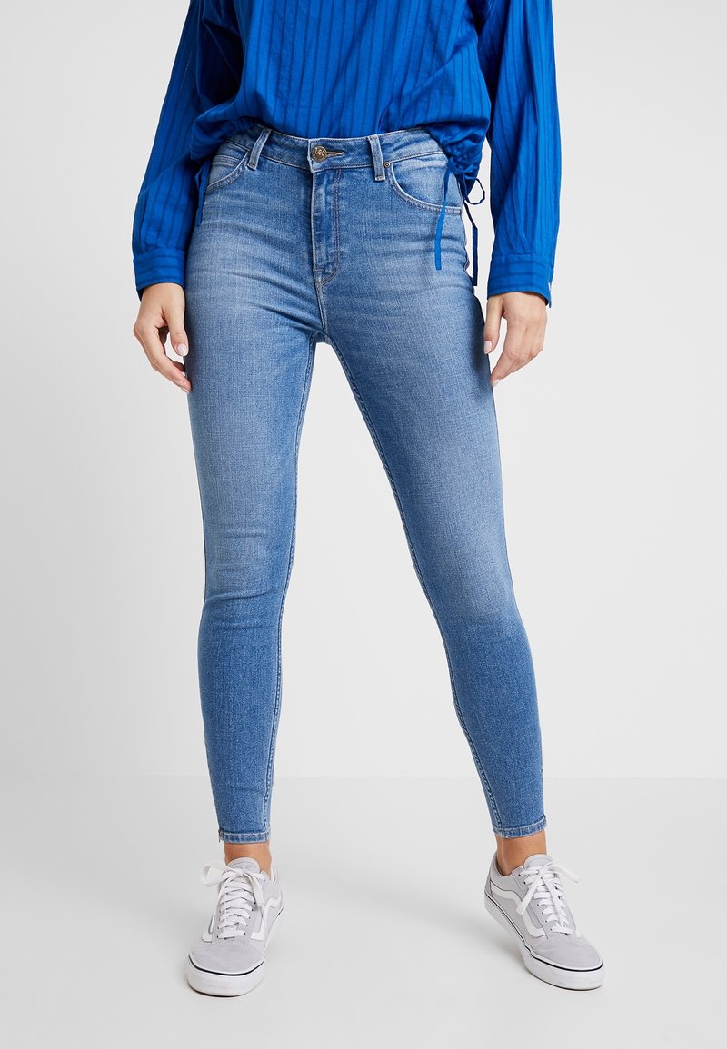 Jeans ajustados de mezclilla en azul, con una cintura de altura media, que cuentan con un cierre de botón, bolsillos delanteros y desvanecimiento mínimo, combinados con zapatillas gris.