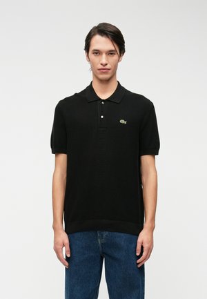 Polo shirt nero con un tessuto a maglia testurizzato, maniche corte e colletto. Presenta un piccolo logo verde Lacoste sopra la tasca sinistra del petto.