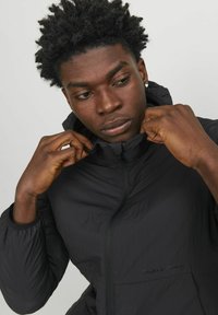 Black hooded jacket with a smooth, lightweight texture. Features a zipper closure and logo detail on the front. Model adjusts the collar.