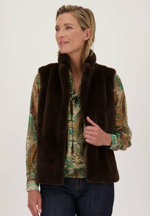 Gilet en fausse fourrure marron foncé avec un col haut, présentant une texture douce et un design à ouverture sur le devant, porté sur une blouse à motifs et un jean foncé.