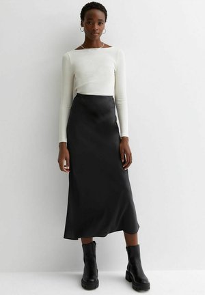 Weißes Langarmoberteil kombiniert mit einem schwarzen Satin-Midirock und schwarzen klobigen Stiefeletten. Schlichte Silhouette mit glatter Textur.