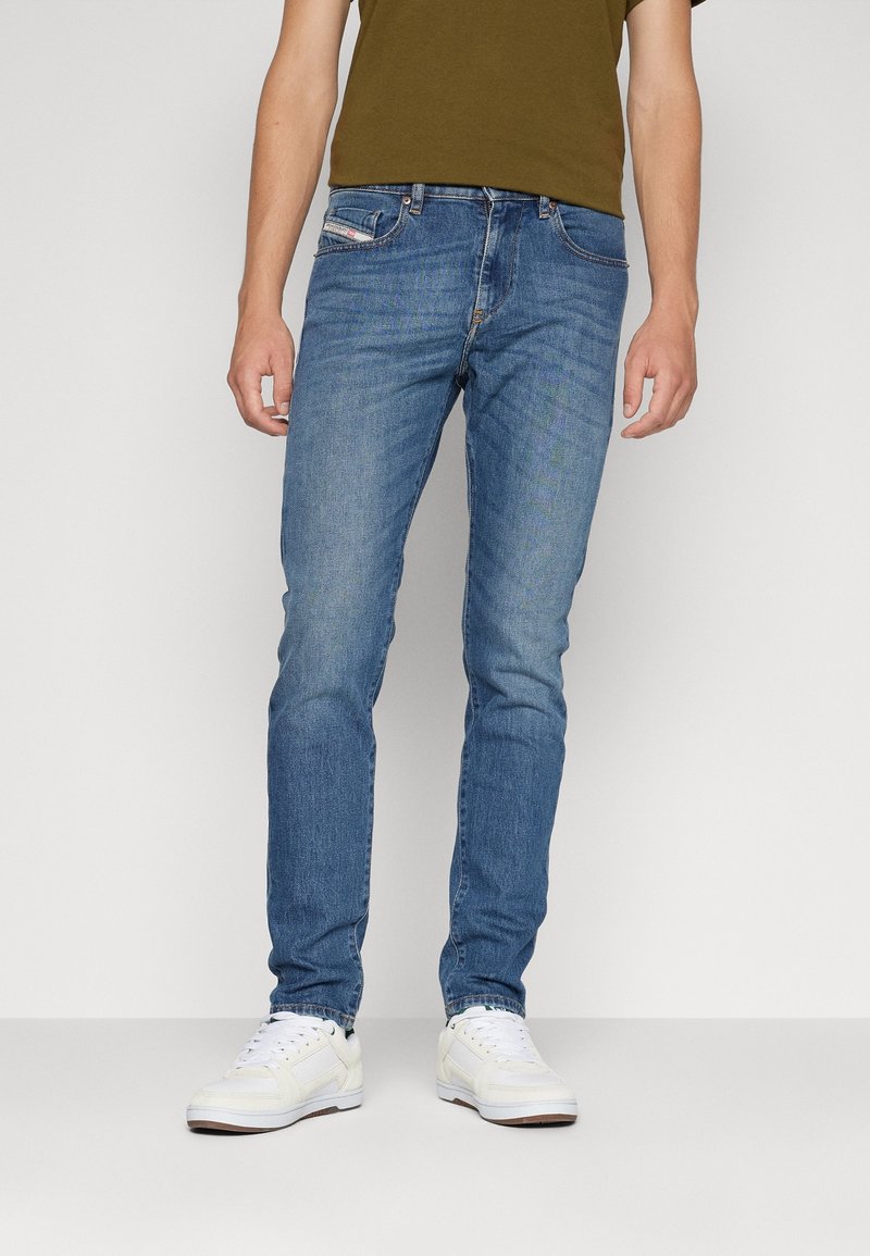 Muž v modrých slim-fit džínách, olivově zeleném tričku a bílých teniskách stojící na světlém jednobarevném pozadí.