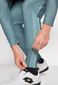 Lys teal treningsbukser med slim fit, som har en sidelommer med glidelås og hvite detaljer, kombinert med hvite sko og svarte sokker.