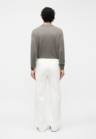 Homme aux cheveux bouclés et foncés, vu de dos, portant une chemise grise à manches longues rentrée dans un pantalon blanc à jambes larges, sur un fond uni.