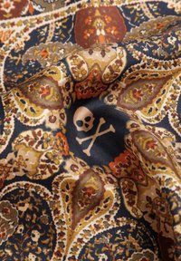 Tela negra con intrincados patrones de paisley en dorado, naranja y marrón, con un diseño central de una calavera y huesos cruzados en blanco.