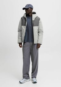 Gepolsterte Jacke in Hellgrau mit dunkelgrauen Akzenten, kombiniert mit einem blauen T-Shirt und grauen Hosen. Verfügt über eine Kapuze und Reißverschlusstaschen.