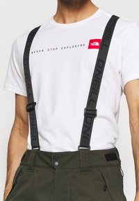 Vit t-shirt med texten "NEVER STOP EXPLORING" och röd logotyp, i kombination med olivgröna byxor med svarta hängslen och tryckknappsstängning.