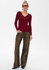 Bordowy prążkowany sweter z długim rękawem i dekoltem w serek w połączeniu z szerokimi spodniami w leopardzi nadruk. Modelka nosi masywne okulary przeciwsłoneczne i trzyma jedną rękę w kieszeni.
