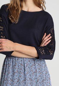 Marineblå topp med spetsdetaljer och långa ärmar, i kombination med en ljusblå blommig kjol. Har en rund halsringning och avslappnad passform.