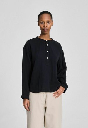 Vrouw met natuurlijk haar naar achteren gebonden, gekleed in een zwart, lang mouws, geknoopt overhemd en beige broek met wijde pijpen, staand tegen een effen grijze achtergrond.