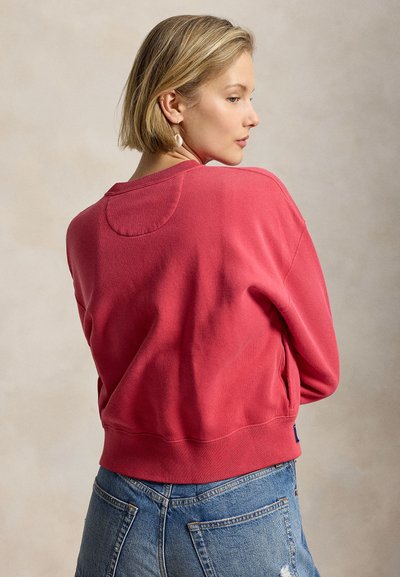 Γυναίκα με κοντά ξανθά μαλλιά που φοράει κοραλλί ροζ πουλόβερ και μπλε τζιν, κοιτάζοντας πλάγια μπροστά σε ανοιχτό μπεζ φόντο.