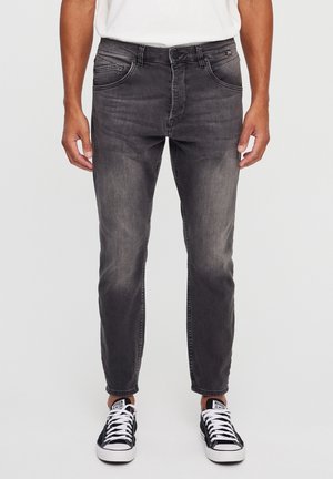 Mand iført falmede sorte slim-fit jeans og sort-hvide lave sneakers, stående mod en ensfarvet baggrund.