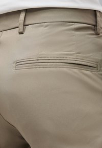 Detail béžových nohavíc so zadným vreckom s horizontálnym rezom a pútkami na opasok na páse.