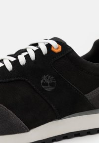 La sneaker en suède noir et maille présente des lacets blancs, un accent orange et un logo subtil. La semelle est blanche avec un motif de gomme noir texturé.