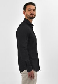 Zwarte langemouwen shirt met een geknoopte kraag, slim fit, gladde textuur. Gepaard met lichtgekleurde broek, model staat zijwaarts.