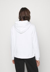Felpa bianca realizzata in tessuto morbido, con un grande cappuccio e polsini a coste, con orlo dritto. Indossata con pantaloni neri.