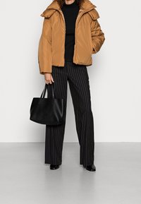 Puffatakki lämpimässä ruskeassa värissä, korkealla kauluksella, yhdistettynä mustiin raidallisiin leveälahkeisiin housuihin ja mustaan kassi, jossa on teksturoituja yksityiskohtia.