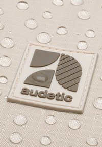 Beige szövet kiemelkedő pöttyökkel és cseppekkel, barna és krémszínű domború logóval. A logó geometrikus formákat és szöveget tartalmaz.