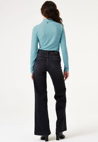 Türkiser Langarm-Top mit strukturiertem Muster, kombiniert mit hochgeschnittenen schwarzen Jeans. Das Outfit hat eine taillierte Passform und ausgestellte Beine.
