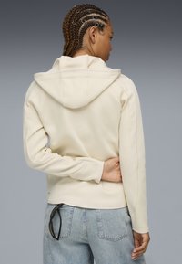 Helles cremefarbenes Sweatshirt mit Kapuze, gerippten Akzenten und einer figurbetonten Passform. Kombiniert mit hellblauen Jeans und einer Sonnenbrille, die hinten gesteckt ist.