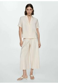 Camicia a maniche corte color crema con bottoni abbinata a pantaloni a gamba larga, entrambi in un tessuto leggero. Il modello indossa sandali chiari.