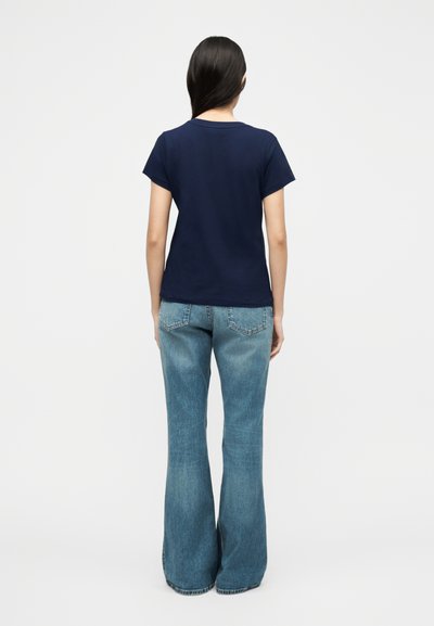 Γυναίκα με μακριά σκούρα μαλλιά, που φοράει μπλε κοντομάνικη μπλούζα και ανοιχτό μπλε τζιν καμπάνα, στέκεται με την πλάτη προς το μέρος μας μπροστά σε λευκό φόντο.