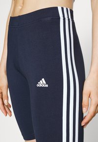 Marineblauwe fietsshorts van rekbaar materiaal met een wit driekleurig ontwerp langs de zijkanten en een Adidas-logo op de dij.