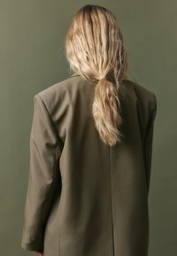 Olive roheline ülemõõduline bleiser struktureeritud õlgadega, sile kangas ja üks tagumine lõhik; pikad, lainelised juuksed voogavad õlgadele.