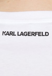 Camiseta de algodón blanca con un logo negro de "KARL LAGERFELD" impreso en la parte posterior, cerca del cuello. Textura suave y ligera.