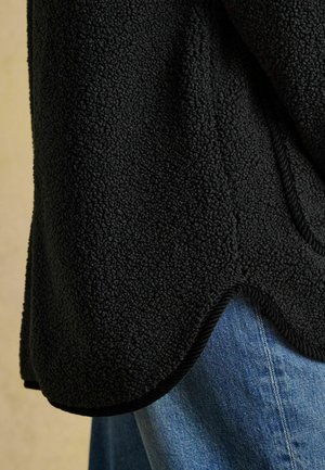 Schwarze Fleecejacke mit strukturierter Oberfläche, abgerundetem Saum und kontrastierendem schwarzen Besatz, kombiniert mit hellblauem Denim-Stoff.