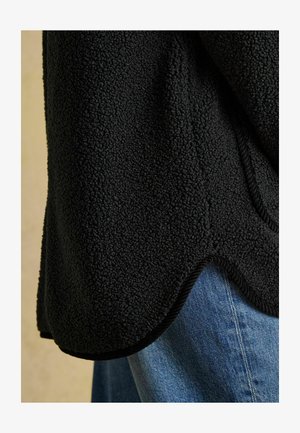 Schwarze Fleecejacke mit strukturierter Oberfläche, abgerundetem Saum und kontrastierendem schwarzen Besatz, kombiniert mit hellblauem Denim-Stoff.