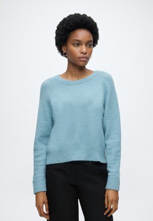 Femme aux cheveux courts et bouclés, portant des boucles d'oreilles en anneaux dorés, un pull bleu clair à manches longues et un pantalon noir, debout devant un fond uni.