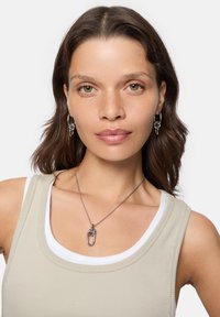 Söljtonad lagerhalsband med ett framträdande hängande paperclip-formad berlock, bärs över en ljusbeige linne med vita detaljer.