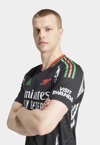 Maillot de sport noir à manches courtes, avec des accents verts et rouges. Présente les logos "Emirates Fly Better" et "Visit Rwanda". Matériau texturé visible.