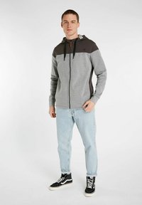 Gray zip-up hoodie with a black upper section, adjustable hood, and drawstrings, worn over light blue jeans and black high-top sneakers.