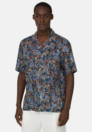 Homme portant une chemise à manches courtes à boutons avec un imprimé floral bleu et marron, et un pantalon blanc, debout devant un fond clair uni.